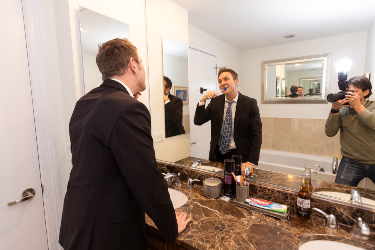 Jon brushes his teeth in a suit while a photographer takes his picture.